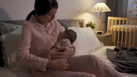 cropped shot of asian new mother sitting on bed is patting and coaxing her wailing baby at night during bedtime in the bedroom at home.