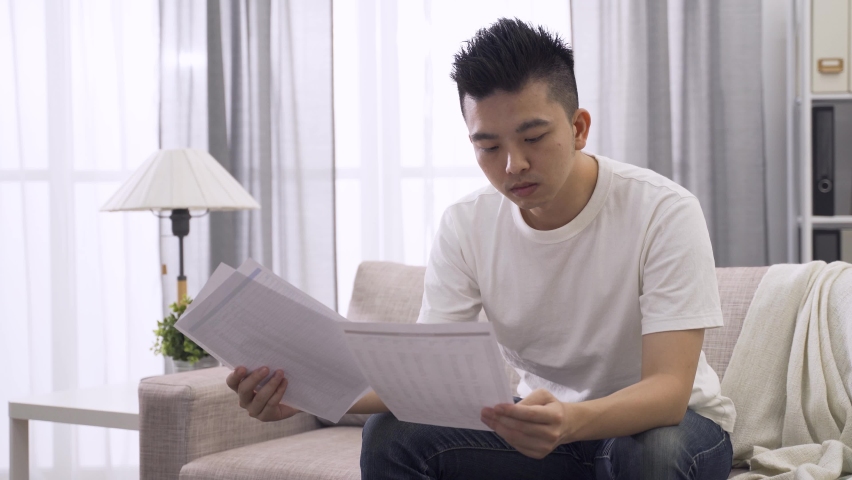 College student holding comparing and referring to reading materials for his paper in the living room. with great concentration. | Shutterstock HD Video #1060191419