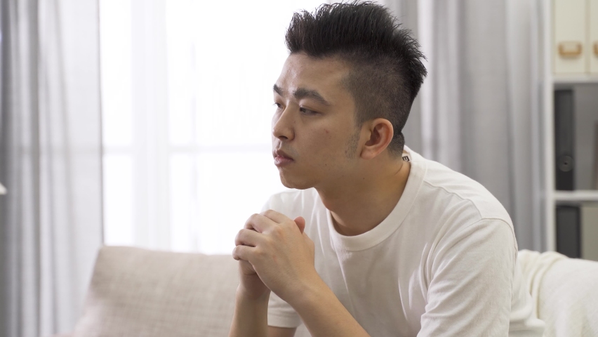 Headshot against bright background chinese male lost in thoughts is pondering with chin on his clasped hands | Shutterstock HD Video #1060191422