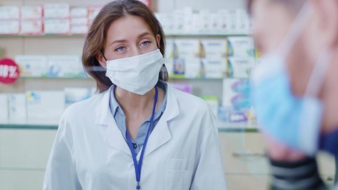 Female apothecarist working in drugstore consulting customers. Careful smart woman pharmacist listening to symptoms wearing protective mask selling drugs.