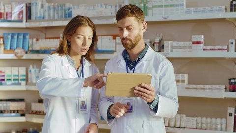 Two caucasian pharmacists using tablet computer negotiating reading medical treatment details cooperating in the pharmacy. Drugs store. Apothecary.