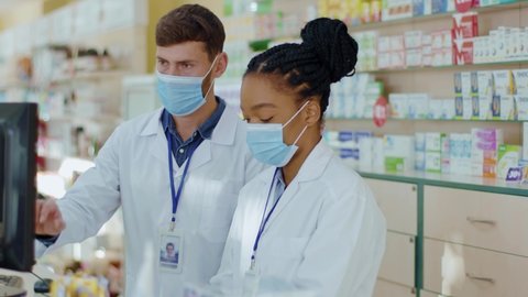 Multi-ethnic couple of pharmacy experts wearing protective masks coworking in drugstore during coronavirus pandemic. Quarantine. Pharmacy. Medications.