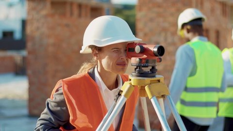 Close up young woman surveyor in work clothes and helmet takes measures with theodolite stand helmet hardhat geodesy digital engineering real estate teamwork infrastructure industry slow motion