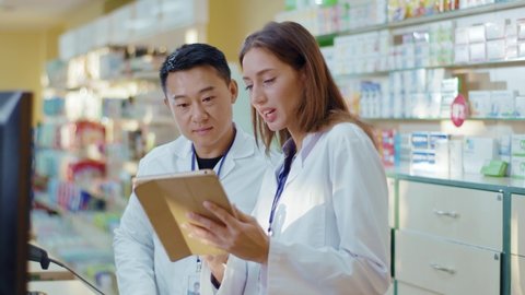Asian adult and younger female intern cooperating in drugstore. Aspiring woman apothecarist asking advice from male colleague learning drugs in pharmacy using tablet.