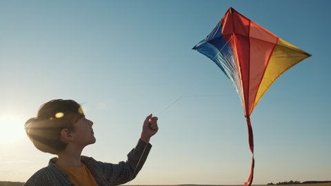 Happy boy launches bright kite into sky on mown wheat field, playing with wind in field of an orange sunset on day lens flares wind turbines in summer slow motion. School break. Lifestyle. Childhood