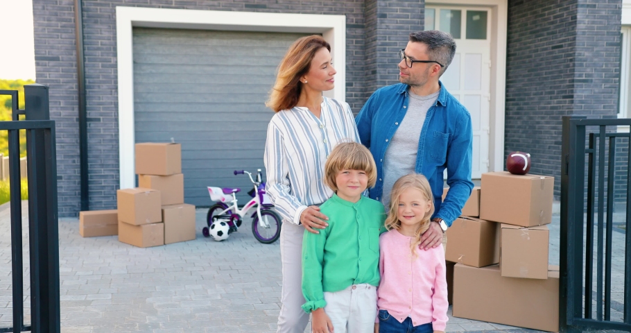 Portrait of happy Caucasian family with small children at new house in suburb smiling. Carton boxes on background. Cheerful parents and kids moving in new home. Settlement in outskirt. | Shutterstock HD Video #1060198871