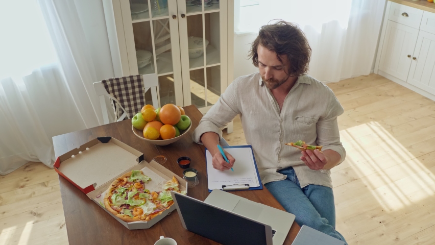 Attractive Student Studying Remotely With A Laptop From Home. Man Sitting at the Table Eat a Pizza During Remotely Study From Home. Young Man With Bristle Sitting at Bright Cozy Kitchen. Royalty-Free Stock Footage #1060198982