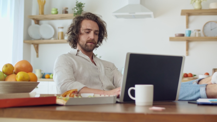 Attractive Tired Man Working With A Laptop From Home. Man Sitting at the Table Eat a Pizza During Remotely Work From Home. Young Man With Bristle Sitting and Put His Legs on the Table at Cozy Kitchen. Royalty-Free Stock Footage #1060198988