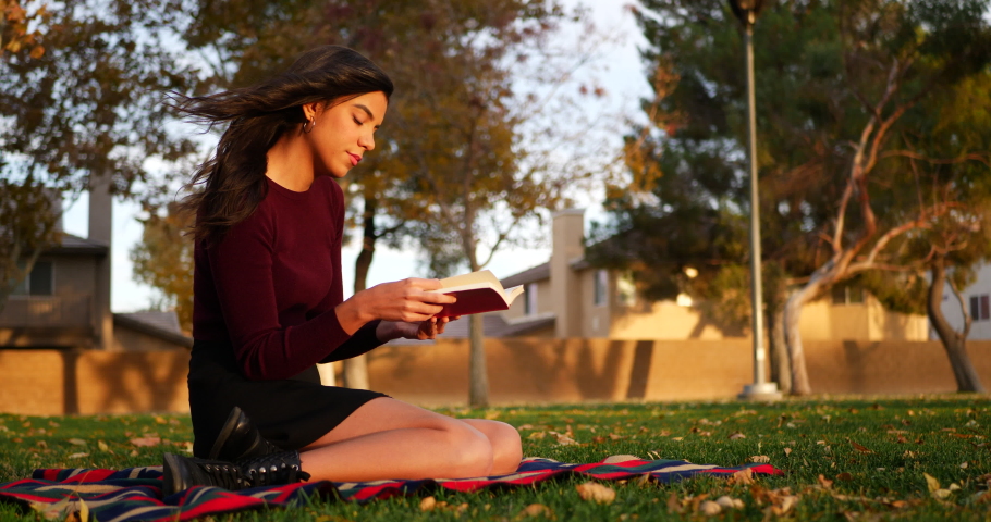 A gorgeous young hispanic woman student reading a book or novel outdoors on a college university campus SLIDE LEFT.