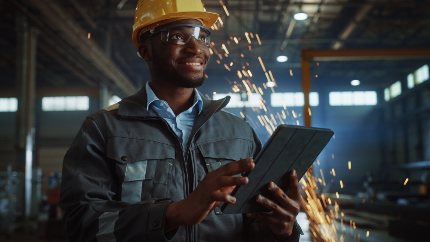Professional Heavy Industry Engineer/Worker Wearing Safety Uniform and Hard Hat Uses Tablet Computer. Smiling African American Industrial Specialist Standing in a Metal Construction Manufacture. | Shutterstock HD Video #1060203755