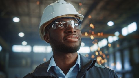 Happy Professional Heavy Industry Engineer/Worker Wearing Uniform, Glasses and Hard Hat in a Steel Factory. Smiling African American Industrial Specialist Standing in a Metal Construction Manufacture.