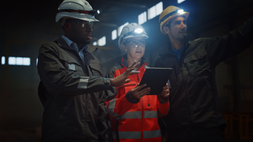 Three Diverse Multicultural Heavy Industry Engineers and Workers in Uniform Stand in Dark Steel Factory Using Flashlights on Their Hard Hats. Female Industrial Contractor is Using a Tablet Computer. Royalty-Free Stock Footage #1060203830