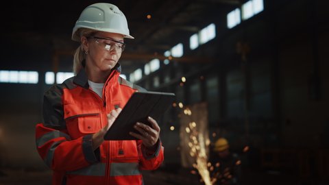 Professional Heavy Industry Engineer/Worker Wearing Safety Uniform and Hard Hat Uses Tablet Computer. Serious Successful Female Industrial Specialist Standing in a Metal Manufacture Warehouse.
