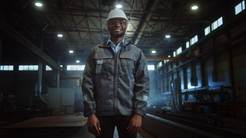 Happy Professional Heavy Industry Engineer/Worker Wearing Uniform, Glasses and Hard Hat in a Steel Factory. Smiling African American Industrial Specialist Standing in a Metal Construction Manufacture. | Shutterstock HD Video #1060203869