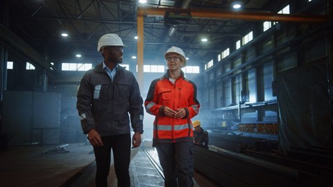 Two Heavy Industry Engineers in Hard Hats Walk in Steel Metal Manufacturing Factory and Have a Discussion. Black African American Industrial Specialist Talk to Female Technician in Manufacture.