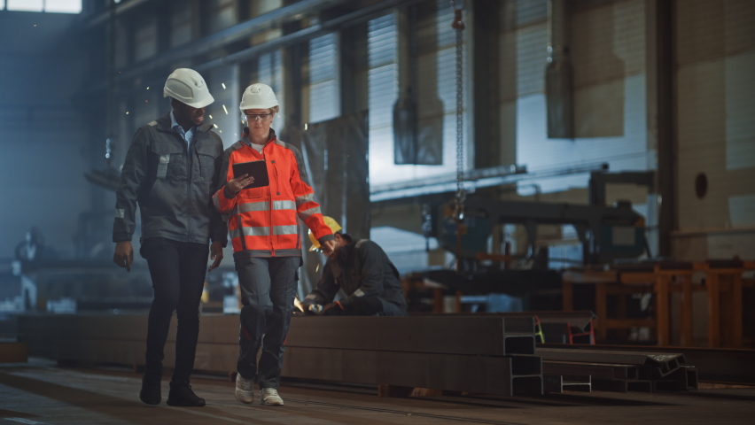 Two Heavy Industry Engineers Walk in Steel Factory, Use Tablet and Discuss Work. Industrial Worker Uses Angle Grinder in the Background. Black African American Specialist Talks to Female Technician. Royalty-Free Stock Footage #1060203881