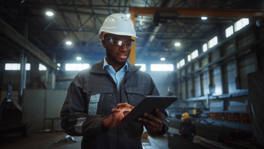 Professional Heavy Industry Engineer/Worker Wearing Safety Uniform and Hard Hat Uses Tablet Computer. Smiling African American Industrial Specialist Walking in a Metal Construction Manufacture. | Shutterstock HD Video #1060203884