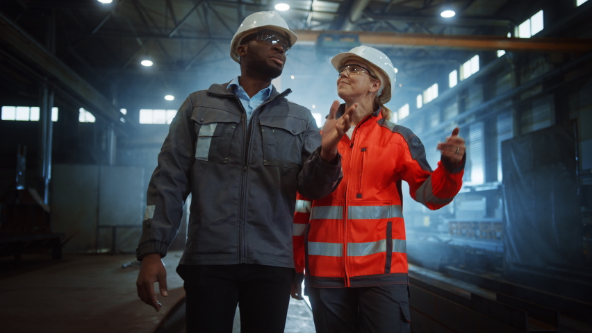 Two Heavy Industry Engineers in Hard Hats Walk in Steel Metal Manufacturing Factory and Have a Discussion. Black African American Industrial Specialist Talk to Female Technician in Manufacture. | Shutterstock HD Video #1060203887