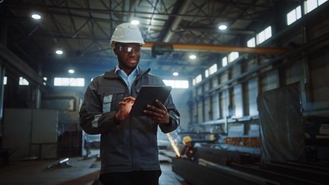 Professional Heavy Industry Engineer/Worker Wearing Safety Uniform and Hard Hat Uses Tablet Computer. Smiling African American Industrial Specialist Walking in a Metal Construction Manufacture.