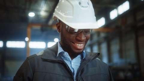 Professional Heavy Industry Engineer/Worker Wearing Safety Uniform and Hard Hat Uses Tablet Computer. Smiling African American Industrial Specialist Walking in a Metal Construction Manufacture.