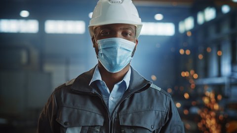 Professional Heavy Industry Engineer/Worker Wearing Safety Face Mask, Uniform and Hard Hat in a Steel Factory. African American Industrial Specialist Standing in a Metal Construction Manufacture.