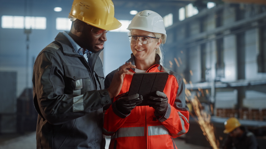 Two Heavy Industry Engineers Stand in Steel Metal Manufacturing Factory, Use Digital Tablet Computer and Have a Discussion. Black African American Industrial Specialist Talk to Female Technician. | Shutterstock HD Video #1060203962