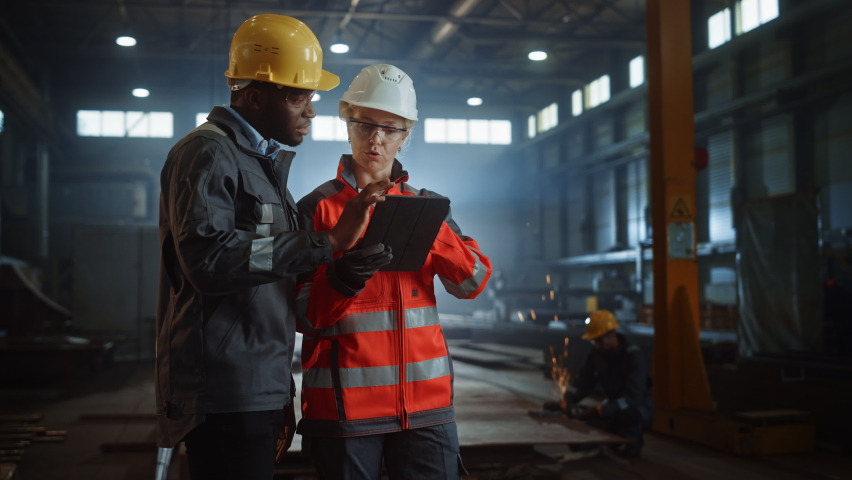 Two Heavy Industry Engineers Stand in Steel Metal Manufacturing Factory, Use Digital Tablet Computer and Have a Discussion. Black African American Industrial Specialist Talk to Female Technician. | Shutterstock HD Video #1060203974