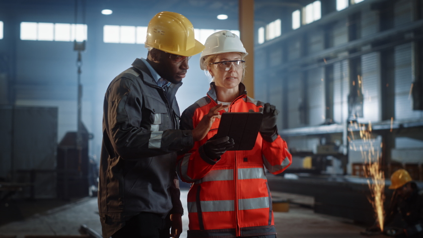Two Heavy Industry Engineers Stand in Steel Metal Manufacturing Factory, Use Digital Tablet Computer and Have a Discussion. Black African American Industrial Specialist Talk to Female Technician. | Shutterstock HD Video #1060203983
