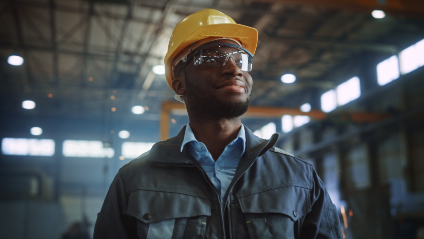 Happy Professional Heavy Industry Engineer/Worker Wearing Uniform, Glasses and Hard Hat in a Steel Factory. Smiling African American Industrial Specialist Standing in a Metal Construction Manufacture. | Shutterstock HD Video #1060203989