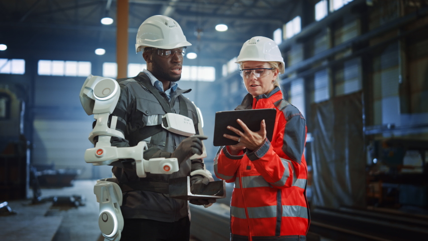 Futuristic Concept of a Manual Labor Worker in a Bionic Exoskeleton Prototype Working in a Factory. Heavy Industry Engineer Monitors the Powered Suit on an African American Assistant. | Shutterstock HD Video #1060204016