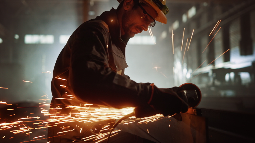 Heavy Industry Engineering Factory Interior with Industrial Worker Using Angle Grinder and Cutting a Metal Tube. Contractor in Safety Uniform and Hard Hat Manufacturing Metal Structures. Royalty-Free Stock Footage #1060204070