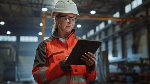 Professional Heavy Industry Engineer/Worker Wearing Safety Uniform and Hard Hat Uses Tablet Computer. Serious Successful Female Industrial Specialist Walking in a Metal Manufacture Warehouse.