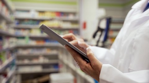 Close up of female pharmacist scrolling through prescriptions on digital tablet standing in white coat in clean pharmacy 