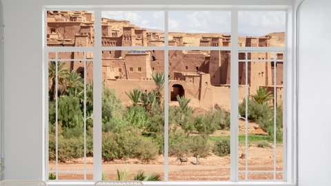 Ait Ben Haddou cityscape as seen from a white window while birds are flying. It is seen Kasbah near Ouarzazate in the Atlas Mountains of Morocco. UNESCO World Heritage Site since 1987. The red village