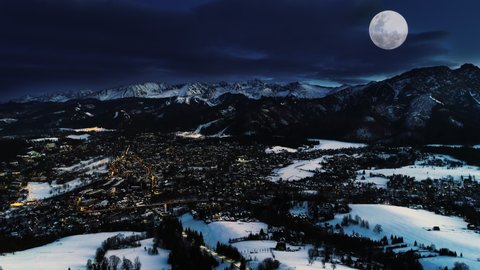 Mountains at winter night, aerial view. Magical winter, Christmas village landscape. Illuminated alpine city covered with snow, white mountains, ski resorts and big shining moon. Zakopane at night