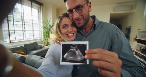 Authentic shot of happy smiling married couple is making video technology call to friends or family with smartphone from home and showing an ultrasound to announce their future child birth.