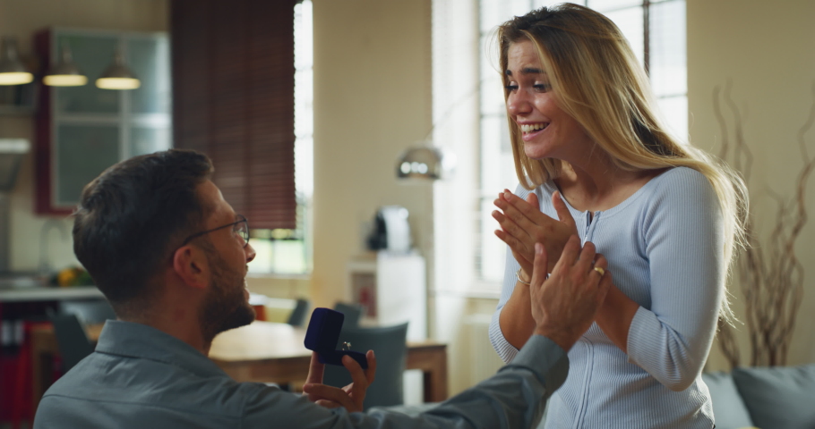 Authentic shot of young handsome man is making a surprise proposal of marriage to his beloved woman in living room at home. The woman is accepting emotionally and giving a hug full of love