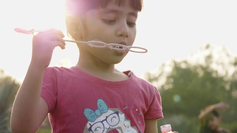 A close-up shot of a cute and adorable little girl child is blowing soap water bubbles against the sun in the park or garden. 