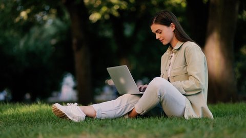 Busy attractive woman working at the laptop as sitting on grass in city park. Student studying outdoor. Freelancer working in the park