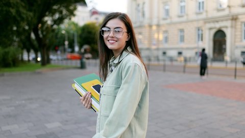 Student girl with books walking to the university building. Happy and smiling, looking at the camera and walking forward.
