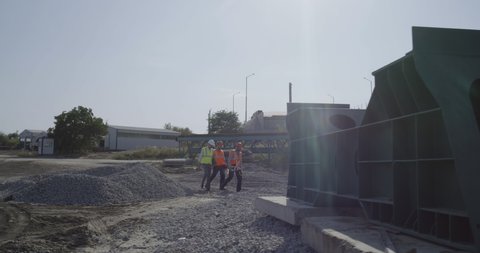 Tracking shot of group of male contractors in hardhats and waistcoats walking on construction site and talking on sunny day