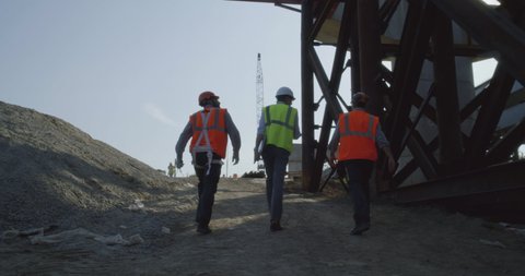 Tracking shot of men in vests and helmets walking on path and talking during inspection on bridge construction site