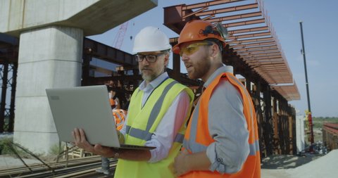 Mature and adult men in uniform browsing and discussing blueprint on laptop while working on construction site together