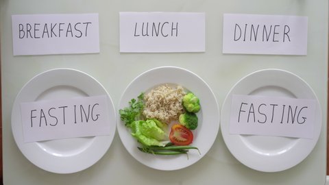 Hands of a woman put a plate with brown rice and vegetables under the title lunch and under the title breakfast and dinner she puts empty plates with titles fasting. Interval fasting concept. Skipping