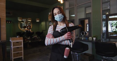 Portrait of Caucasian female hairdresser working in hair salon wearing face mask with hair dryer, crossing her arms, slow motion. Health and hygiene in workplace during Coronavirus Covid 19 pandemic.