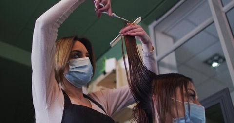 Caucasian female hairdresser working in hair salon wearing face mask, cutting hair of Caucasian woman in face mask, slow motion. Health and hygiene in workplace during Coronavirus Covid 19 pandemic.