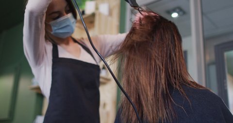 Caucasian female hairdresser working in hair salon wearing face mask, drying hair of female Caucasian customer, slow motion. Health and hygiene in workplace during Coronavirus Covid 19 pandemic.