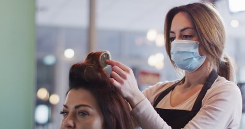 Caucasian female hairdresser working in hair salon wearing face mask, putting hair roller on hair of Caucasian woman, slow motion. Health and hygiene in workplace during Coronavirus Covid 19 pandemic.