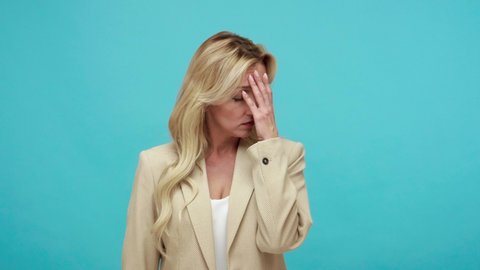 Exhausted tired adult woman with blond hair doing facepalm gesture, shocked and depressed with stupid situation, ashamed with mistake, loser. Indoor studio shot isolated on blue background