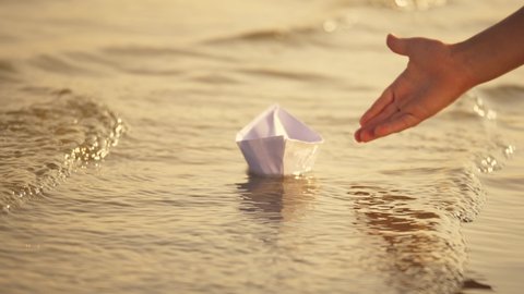 Hand of a little girl lowers a paper boat into the river. Children's paper boat on waves of the river. Close-up of a little girl's hand with a paper boat. Child playing on the river beach at sunset